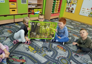 Dzieci siedzą na dywanie. Eksperci pokazują planszę edukacyjną.